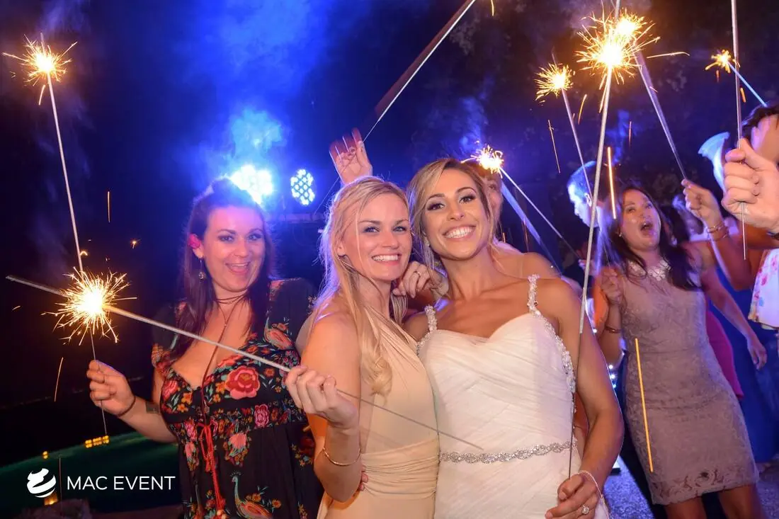 A joyful bride in a white gown is surrounded by friends and family, all holding sparklers, celebrating under vibrant blue lighting at a wedding reception.
