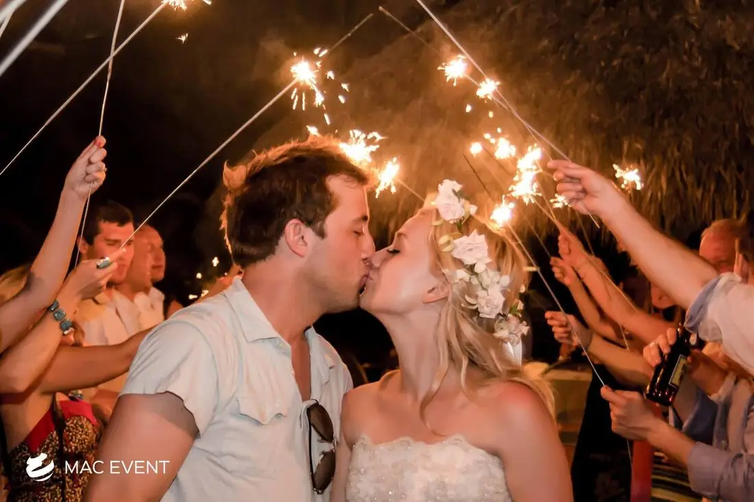 A newlywed couple shares a romantic kiss under a night sky illuminated by sparklers held by their guests, creating a magical wedding atmosphere.